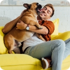Man sits on a yellow couch and hugs a bulldog.